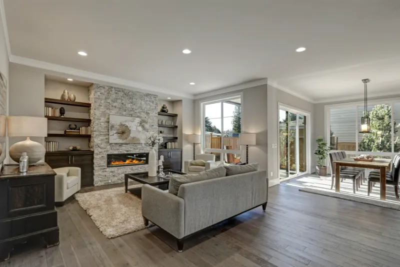 Modern living and dining area with gray sofas, stone fireplace, built-in shelves, large windows, hardwood floors, and a dining table near sliding glass doors leading to a fenced backyard.