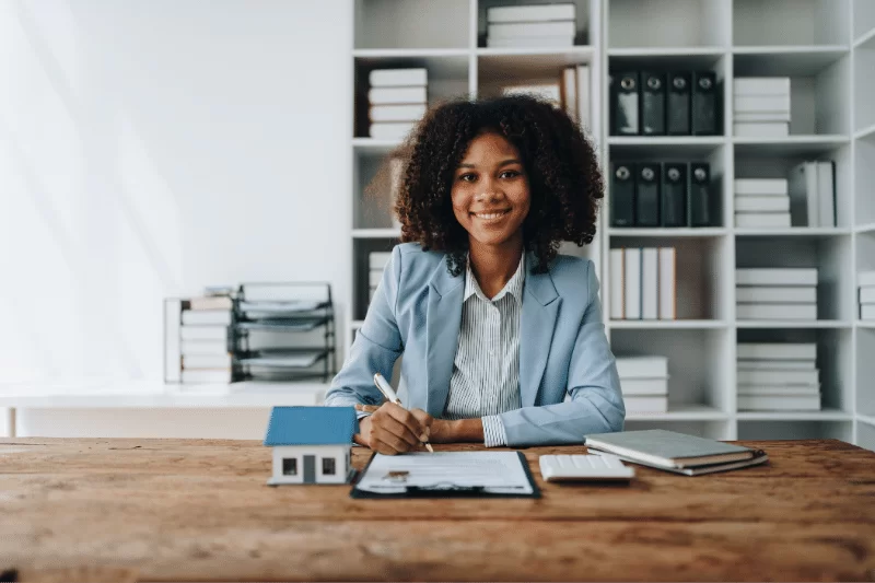 VA Interest Rate Reduction Refinance Loan (IRRRL) 1 A smiling woman in a blazer sits at a desk with paperwork, a model house, a notebook, and a smartphone, in a modern office with shelves of books and binders behind her.